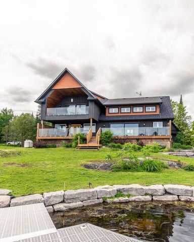 Modern cottage with black and copper exterior, multiple decks, and stone-lined pond with dock in foreground, surrounded by mature trees and manicured lawn.
