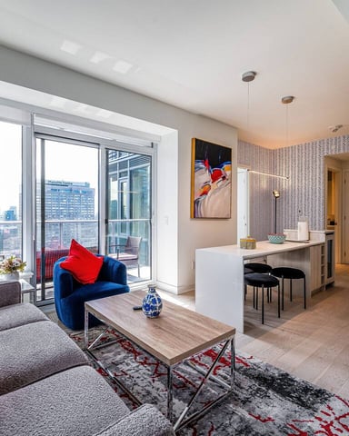 Modern high-rise apartment living room with gray sofa, red pillows, blue armchair, floor-to-ceiling windows overlooking city skyline