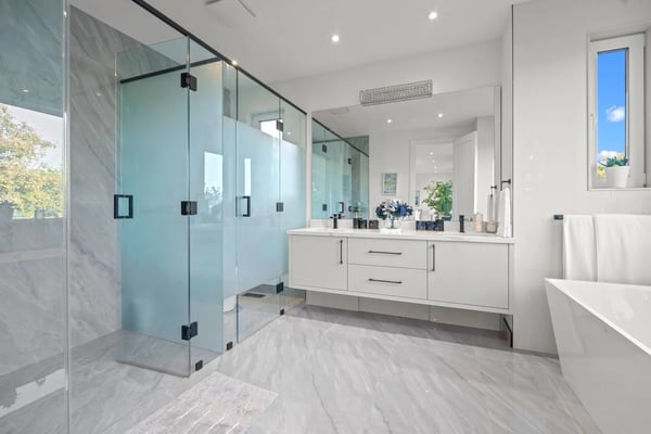 Modern bathroom with marble tile flooring, glass-enclosed shower, white vanity with dual sinks, freestanding tub, and window with blue sky view.