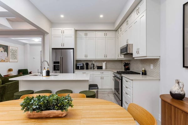 Modern kitchen with white cabinetry, stainless steel appliances, hexagonal tile backsplash, and open-plan dining area with wooden table and green upholstered