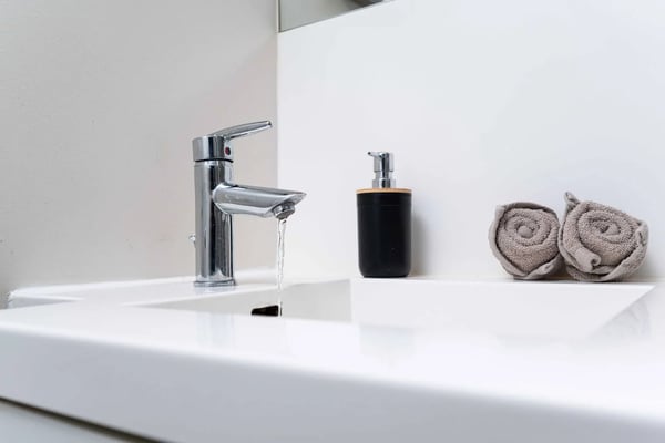 White bathroom sink with chrome faucet running water, black soap dispenser, and rolled gray towels