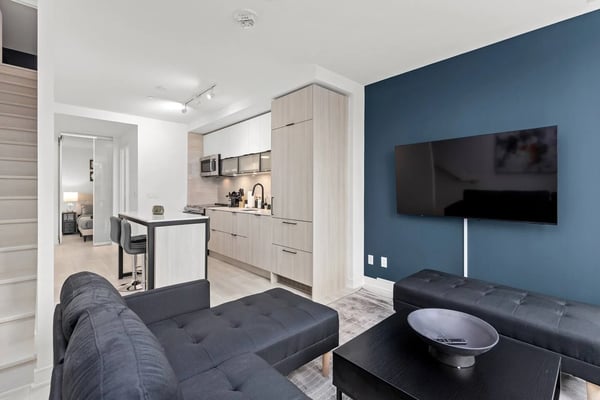 Open-plan studio with dark blue accent wall, black sectional sofa, wall-mounted TV, and light wood kitchen with island seating