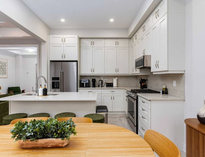 Modern kitchen with white cabinetry, stainless steel appliances, hexagonal tile backsplash, and open-plan dining area with wood table and green upholstered